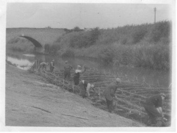 Historic image: a group of labourers using spades and scythes to manage the banks of a large drain. A brick arched bridge is featured in the background.