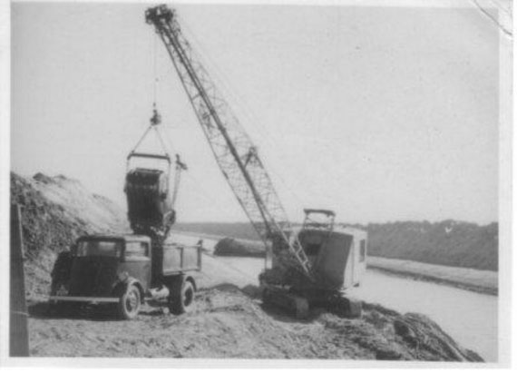 Historic image: an old drag line is pictured, emptying its contents into an old dumper truck which is parked on the lower bank of the drain