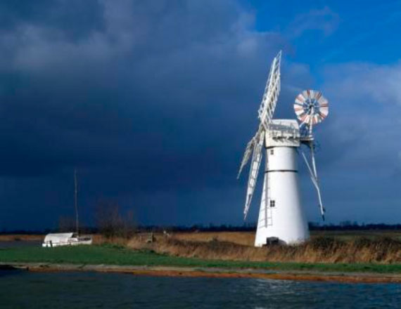 A view of the Thurne Windpump, Thurne Dyke, on the Norfolk Broads, with dark storm clouds gathering over the striking white silhouette of the windmill and it’s sails.