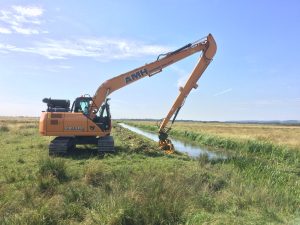 A tracked excavator uses its long-reach hydraulic arm to carry out routine weed cutting and flailing of the drain’s bank sides